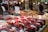 A market stall is filled with containers of fresh strawberries sitting on a wooden counter. Price tags with the number 20 are prominently displayed. A vendor wearing a dark jacket and cap is seen working in the background. The surrounding area includes other produce items and has a bustling atmosphere with various market elements, such as stall signs and lights.