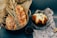 An assortment of freshly baked bread is arranged on decorative paper, including a sesame seed-covered braid loaf and a round loaf. Dried wheat stalks are placed alongside as decor, and a metal scoop with coffee beans adds an additional element. Vintage-style printed paper forms the base, complementing the rustic aesthetic.