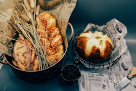 An assortment of freshly baked bread is arranged on decorative paper, including a sesame seed-covered braid loaf and a round loaf. Dried wheat stalks are placed alongside as decor, and a metal scoop with coffee beans adds an additional element. Vintage-style printed paper forms the base, complementing the rustic aesthetic.