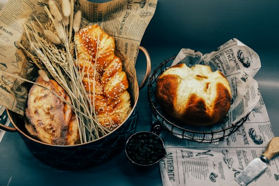 An assortment of freshly baked bread is arranged on decorative paper, including a sesame seed-covered braid loaf and a round loaf. Dried wheat stalks are placed alongside as decor, and a metal scoop with coffee beans adds an additional element. Vintage-style printed paper forms the base, complementing the rustic aesthetic.