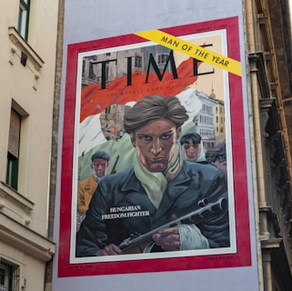 A mural depicting a vintage magazine cover with a central drawing of a stern-faced man in a dark coat and light scarf, labeled 'Hungarian Freedom Fighter.' He holds a weapon, while two other figures stand behind him, one in a yellow coat and another in a blue coat. Buildings and a bright, abstract background fill the backdrop. A yellow banner at the top right corner reads 'Man of the Year.'