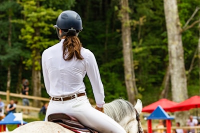 A person wearing a helmet and white riding attire sits on a horse in an outdoor setting. The background features green trees and some blurred red and blue objects, possibly umbrellas or barriers indicating an equestrian event.