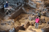 The image depicts three individuals examining a large excavation site containing numerous fossilized remains. One person is standing beside a wooden table with paper, observing the site. The excavation area features deep pits and large skeletal structures partially embedded in the ground.