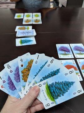 A hand holds several playing cards with tree illustrations and different numbers, set against a dark table. Additional cards are arranged in rows on the table, showing various tree images and numbers.