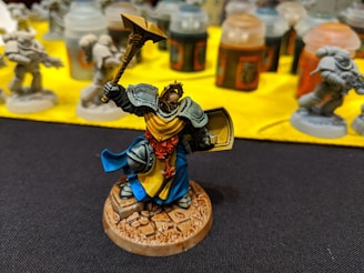 A detailed miniature figure of a warrior with ornate armor, holding a hammer and a shield. The figure has a bright blue and yellow cape and stands on a rocky base. In the background, there are several other unpainted miniatures and paint jars on a yellow surface.