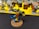 A detailed miniature figure of a warrior with ornate armor, holding a hammer and a shield. The figure has a bright blue and yellow cape and stands on a rocky base. In the background, there are several other unpainted miniatures and paint jars on a yellow surface.
