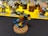 A detailed miniature figure of a warrior with ornate armor, holding a hammer and a shield. The figure has a bright blue and yellow cape and stands on a rocky base. In the background, there are several other unpainted miniatures and paint jars on a yellow surface.