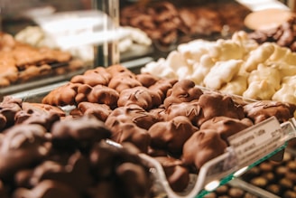 An array of assorted chocolate-covered confections is displayed in a glass case. The focus is on the front row, featuring clusters in various shapes with glossy milk chocolate and white chocolate coatings. The background shows other similar sweet treats, slightly out of focus.