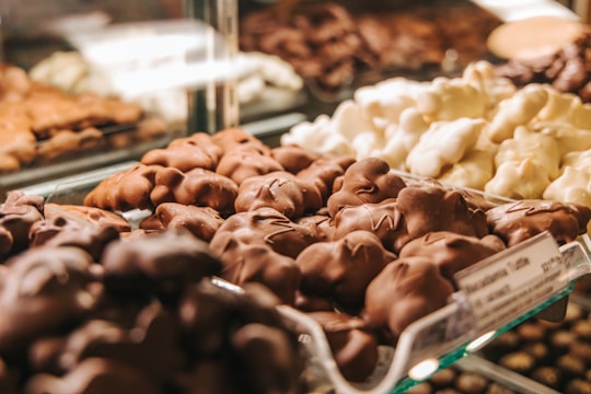 An array of assorted chocolate-covered confections is displayed in a glass case. The focus is on the front row, featuring clusters in various shapes with glossy milk chocolate and white chocolate coatings. The background shows other similar sweet treats, slightly out of focus.