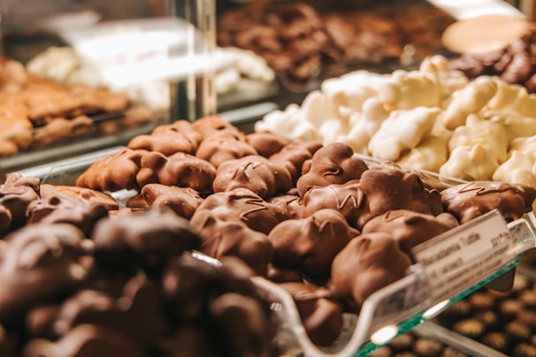 An array of assorted chocolate-covered confections is displayed in a glass case. The focus is on the front row, featuring clusters in various shapes with glossy milk chocolate and white chocolate coatings. The background shows other similar sweet treats, slightly out of focus.