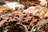 An array of assorted chocolate-covered confections is displayed in a glass case. The focus is on the front row, featuring clusters in various shapes with glossy milk chocolate and white chocolate coatings. The background shows other similar sweet treats, slightly out of focus.