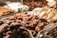 An array of assorted chocolate-covered confections is displayed in a glass case. The focus is on the front row, featuring clusters in various shapes with glossy milk chocolate and white chocolate coatings. The background shows other similar sweet treats, slightly out of focus.