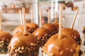 A row of caramel apples is displayed, each coated in a smooth layer of caramel with nuts sprinkled around the bottom. Wooden sticks are inserted into the top of each apple for easy handling. The background is softly blurred, suggesting a setting like a candy shop or market.
