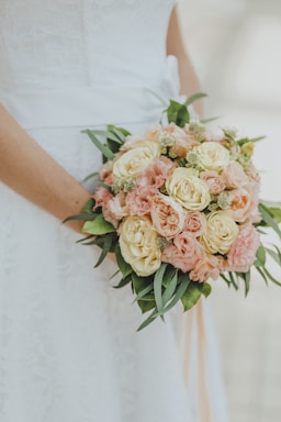 A close-up of a bride holding a bouquet of roses and carnations with hues of pink, peach, and cream, complemented by green foliage. The bride is wearing a lace dress with a ribbon.