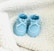 A pair of blue baby shoes with white details resting on a fluffy white blanket. The shoes have small white polka dots and lace-up fronts.
