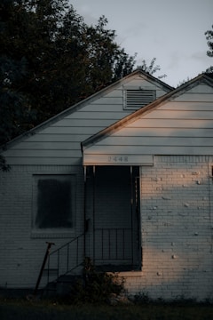 A small, weathered house with white siding and a brick facade. The house number 2448 is visible above the porch, which has a metal railing and overgrown vegetation in front. Soft, warm light illuminates part of the building, adding contrast to the otherwise dim surroundings. Trees with dense foliage surround the house, casting shadows across the roof and exterior walls.