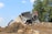 A partially collapsed house amidst a pile of debris being demolished by an excavator in a setting with a clear blue sky and surrounding trees.