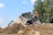 A partially collapsed house amidst a pile of debris being demolished by an excavator in a setting with a clear blue sky and surrounding trees.