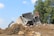 A partially collapsed house amidst a pile of debris being demolished by an excavator in a setting with a clear blue sky and surrounding trees.
