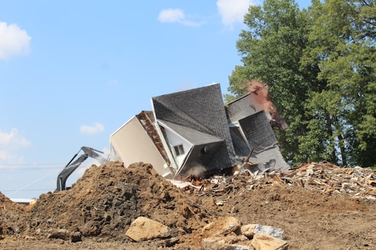 A partially collapsed house amidst a pile of debris being demolished by an excavator in a setting with a clear blue sky and surrounding trees.