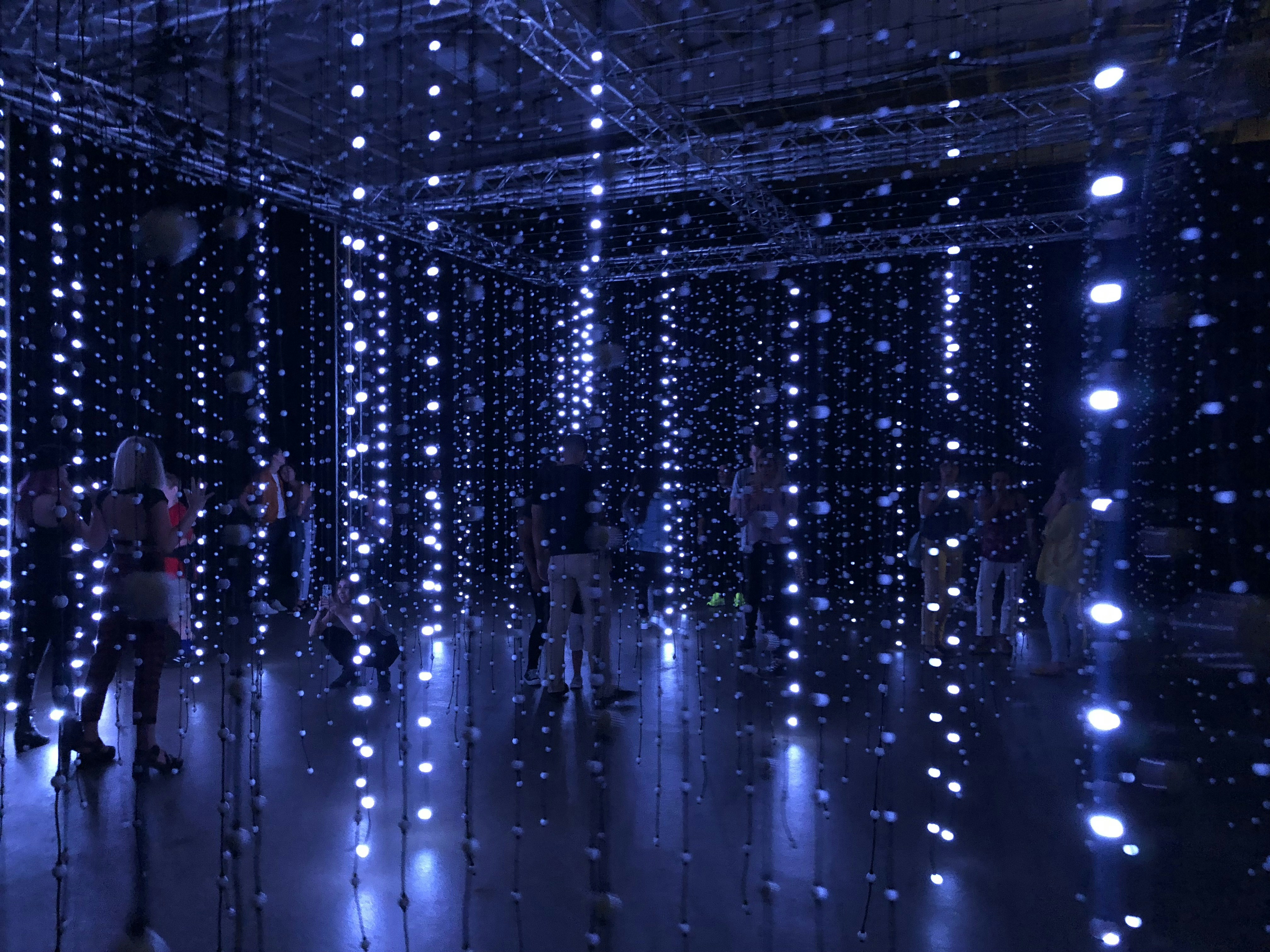 An installation with numerous hanging LED lights in a dark room, creating a glowing, immersive atmosphere. Several people are standing or walking through the installation, interacting with the lights.