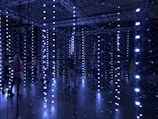 An installation with numerous hanging LED lights in a dark room, creating a glowing, immersive atmosphere. Several people are standing or walking through the installation, interacting with the lights.