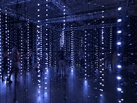 An installation with numerous hanging LED lights in a dark room, creating a glowing, immersive atmosphere. Several people are standing or walking through the installation, interacting with the lights.