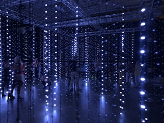 An installation with numerous hanging LED lights in a dark room, creating a glowing, immersive atmosphere. Several people are standing or walking through the installation, interacting with the lights.