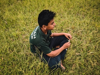 A young person with dark hair sits cross-legged on a field of green grass, wearing a dark green shirt with white text on the sleeve. They appear to be holding something in their hands and are looking forward with a contemplative expression.
