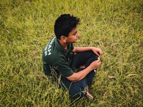 A young person with dark hair sits cross-legged on a field of green grass, wearing a dark green shirt with white text on the sleeve. They appear to be holding something in their hands and are looking forward with a contemplative expression.