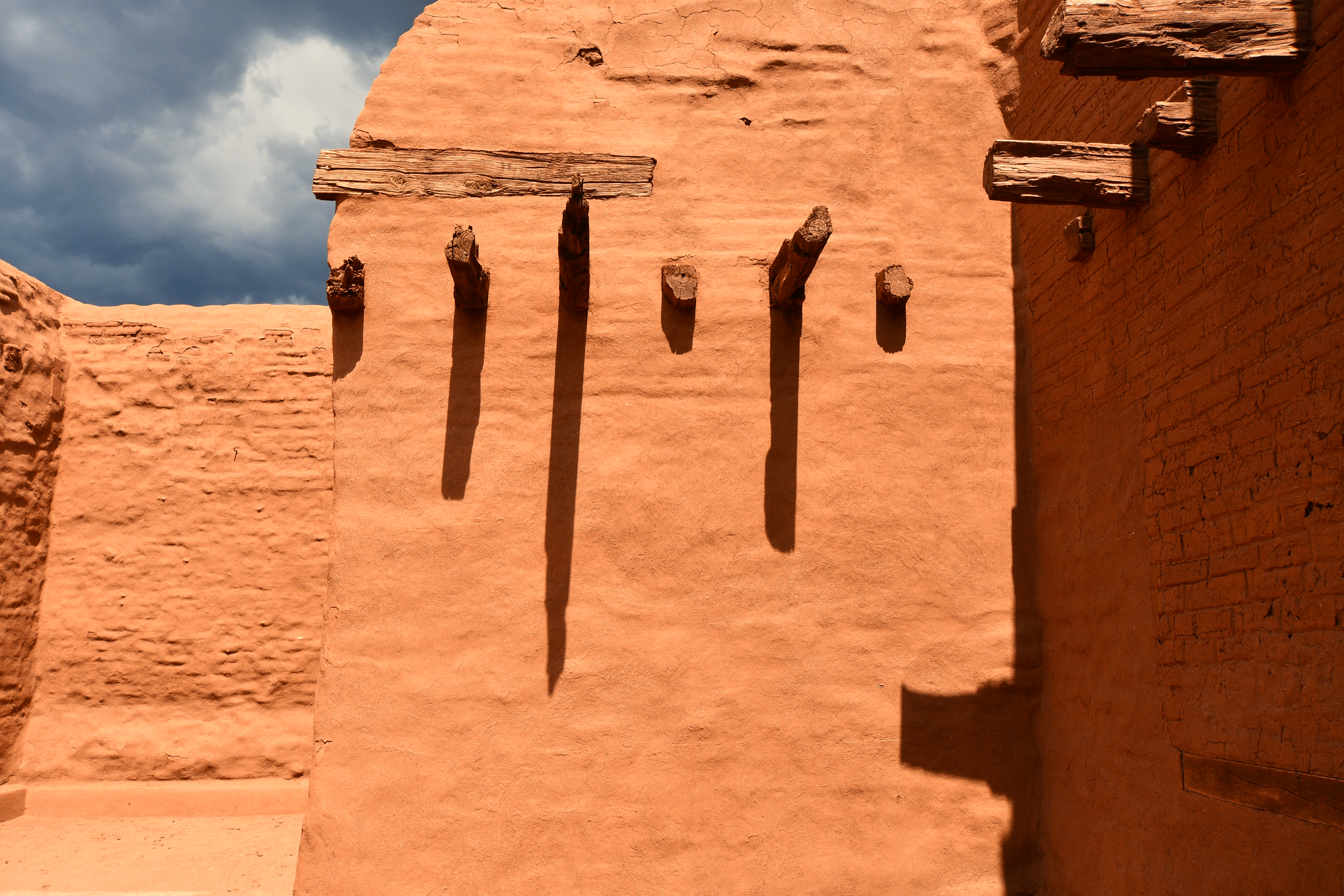brown wooden fence on brown sand, old pueblo style building with long cast shadows from wooden beams