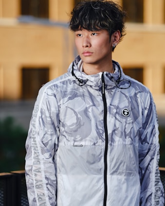 A person is wearing a camouflage-patterned, zip-up hoodie with a design featuring abstract shapes. The hoodie has branding on the chest and sleeves. The background consists of an outdoor urban setting with blurred structures.