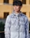 A person is wearing a camouflage-patterned, zip-up hoodie with a design featuring abstract shapes. The hoodie has branding on the chest and sleeves. The background consists of an outdoor urban setting with blurred structures.