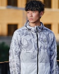 A person is wearing a camouflage-patterned, zip-up hoodie with a design featuring abstract shapes. The hoodie has branding on the chest and sleeves. The background consists of an outdoor urban setting with blurred structures.
