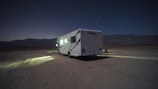 A camper van is parked on a barren, desert-like landscape under a starry night sky. Dim lights from the vehicle cast a small, illuminated area on the ground. Mountain ranges are faintly visible in the background, adding to the secluded atmosphere.