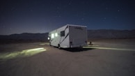 A camper van is parked on a barren, desert-like landscape under a starry night sky. Dim lights from the vehicle cast a small, illuminated area on the ground. Mountain ranges are faintly visible in the background, adding to the secluded atmosphere.