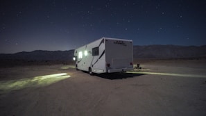 A camper van is parked on a barren, desert-like landscape under a starry night sky. Dim lights from the vehicle cast a small, illuminated area on the ground. Mountain ranges are faintly visible in the background, adding to the secluded atmosphere.