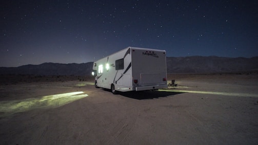 A camper van is parked on a barren, desert-like landscape under a starry night sky. Dim lights from the vehicle cast a small, illuminated area on the ground. Mountain ranges are faintly visible in the background, adding to the secluded atmosphere.
