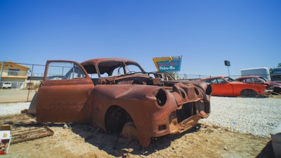 A rusted, abandoned car shell is positioned in a dusty outdoor setting, surrounded by gravel and old car parts. In the background, a sign for a drive-in theater and more derelict vehicles are visible under a clear blue sky.