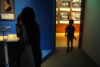In a dimly lit room, a woman and a child are observing displays in what appears to be a museum setting. The child stands in a doorway, silhouetted against a brightly illuminated background featuring historical photographs. The woman is facing an exhibit on the left side that includes a portrait and informational plaques.