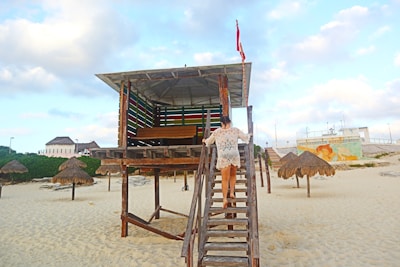 A person in a lacy cover-up walks up a wooden staircase to a lifeguard hut on a sandy beach. The hut is elevated and decorated with horizontal colored slats. There are several thatched umbrellas on the beach, and in the background, buildings and a wall with a mural are visible. The sky is partly cloudy, adding a peaceful setting to the scene.