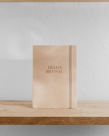 A beige notebook with the words 'HELLO REVIVAL' embossed in gold stands upright on a light wooden shelf against a white textured wall.