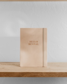 A beige notebook with the words 'HELLO REVIVAL' embossed in gold stands upright on a light wooden shelf against a white textured wall.