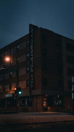 A dimly lit street corner featuring a multi-story brick building with 'The Plainsman' sign. The scene is captured during twilight or early evening, with minimal streetlight illumination and traffic signals glowing red and green. The storefront windows are dark, indicating closed businesses.