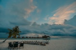 A serene beach scene with an overcast sky, featuring a row of thatched-roof huts on stilts over the water. Palm trees and chairs are positioned along the sandy shore, with people lounging in bean bags nearby, adding a relaxed atmosphere.