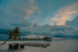 A serene beach scene with an overcast sky, featuring a row of thatched-roof huts on stilts over the water. Palm trees and chairs are positioned along the sandy shore, with people lounging in bean bags nearby, adding a relaxed atmosphere.