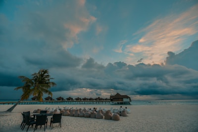 A serene beach scene with an overcast sky, featuring a row of thatched-roof huts on stilts over the water. Palm trees and chairs are positioned along the sandy shore, with people lounging in bean bags nearby, adding a relaxed atmosphere.