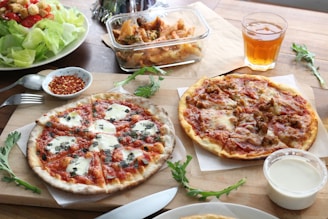 Two pizzas are on a wooden board, one is topped with mozzarella and fresh basil, while the other appears to have sausage and cheese. Nearby, a fresh green salad with tomatoes and croutons is placed in a bowl. A small dish of red pepper flakes is present next to a fork and spoon, with a glass of iced tea and a container of creamy dressing or sauce. In the background, a dish of pasta topped with broccoli is visible.