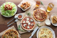 A wooden table is set with a variety of Italian dishes, including several pizzas with different toppings, a fresh green salad with cherry tomatoes and croutons, and a serving of pasta in a glass container. There are small bowls of chili flakes and white dipping sauce, along with slices of toasted bread. Beverages in clear glasses are also visible, likely iced tea and beer. Utensils like a knife and fork are neatly placed nearby.