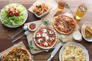 A wooden table is set with a variety of Italian dishes, including several pizzas with different toppings, a fresh green salad with cherry tomatoes and croutons, and a serving of pasta in a glass container. There are small bowls of chili flakes and white dipping sauce, along with slices of toasted bread. Beverages in clear glasses are also visible, likely iced tea and beer. Utensils like a knife and fork are neatly placed nearby.